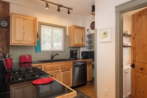 A compact kitchen with wooden cabinets, a black countertop, a red toaster, a sink, a dishwasher, and a window with blinds.