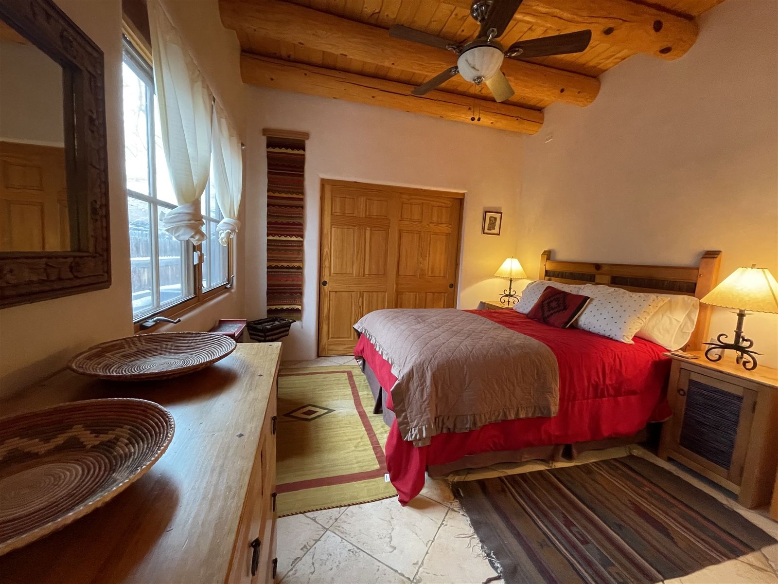 Une chambre à coucher avec un lit, des lampes de chevet, un ventilateur de plafond en bois, une fenêtre avec des rideaux, une porte et une petite table sur laquelle se trouve un panier en osier.