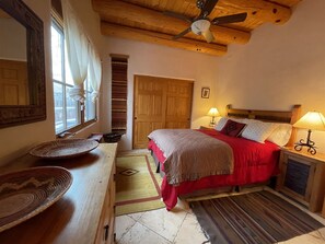 Een slaapkamer met een bed, nachtlampjes, een houten plafondventilator, een raam met gordijnen, een deur en een klein tafeltje met een rieten mand.