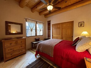 A bedroom with a wooden ceiling, a bed with red bedding, a dresser, a mirror, and a window with curtains.