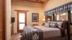 Un dormitorio con cama, puerta, televisor montado en la pared, lámpara y un cuadro enmarcado.