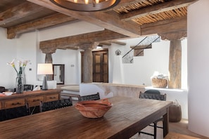 Un interior rústico con vigas de madera a la vista, una mesa de comedor de madera y una escalera con barandilla de hierro forjado.