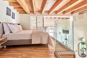 Une chambre avec un plafond en bois, un lit recouvert d’une couvre-lit beige, une table de chevet, une cloison en verre et une plante en pot.