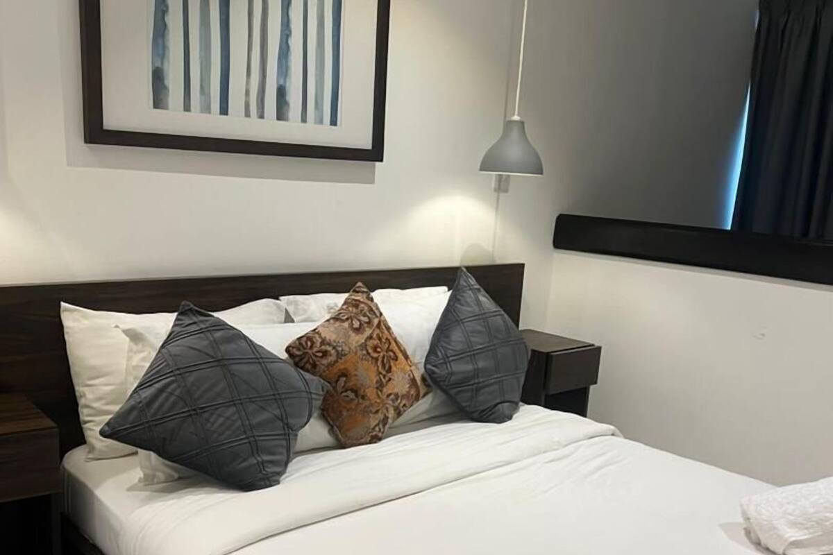 A neatly made bed with white linens and multiple pillows, a framed artwork above the bed, a bedside table with a lamp, and a window with curtains.