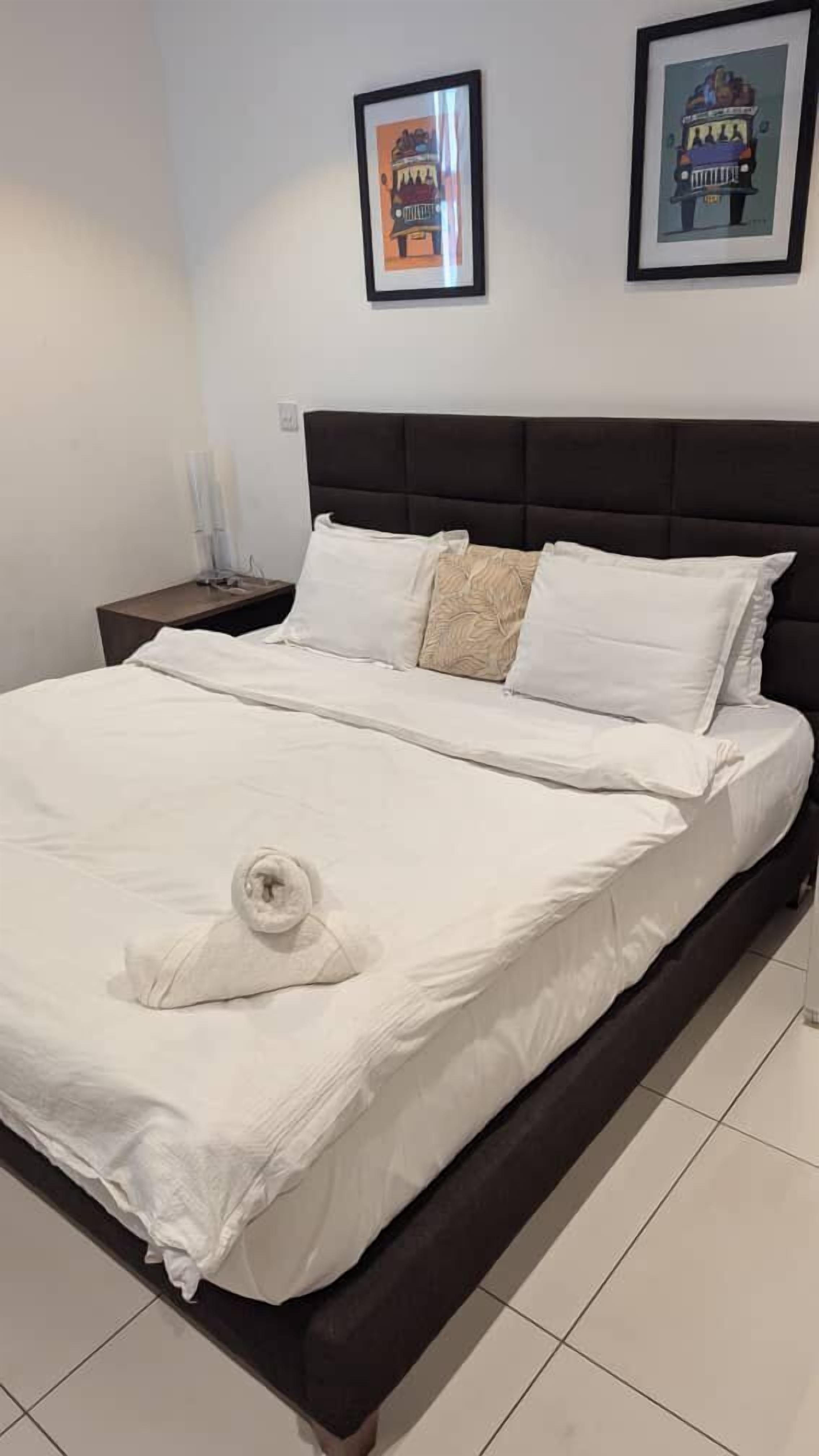A neatly made bed with white linens and a rolled towel, a dark headboard, and two framed artworks on the wall.
