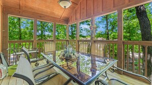 A covered outdoor dining area with a glass table and metal chairs, surrounded by wooden railings and large windows. - Sbl103a -magnolia Moon 1 Bedroom Cabin (Reeds Spring)