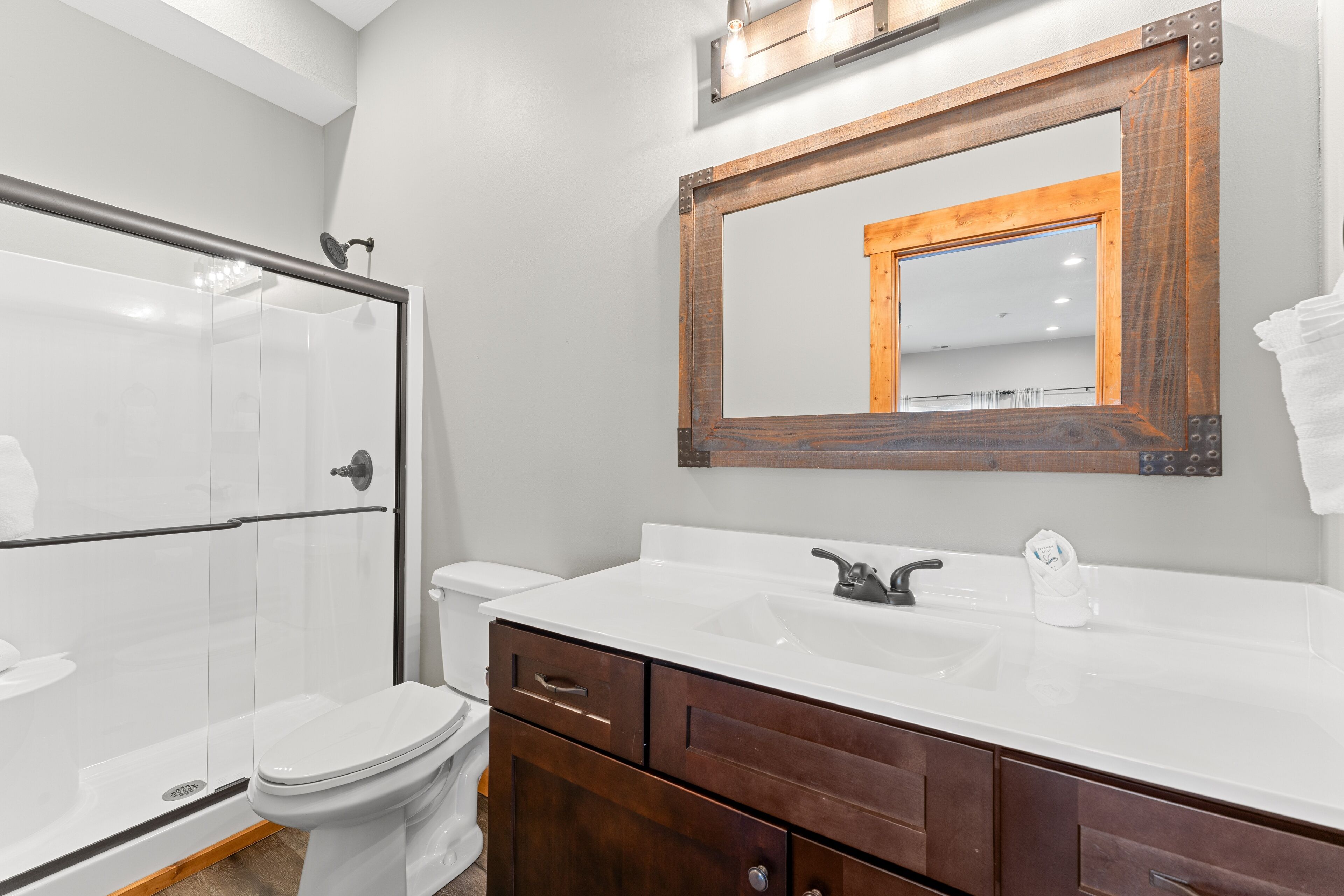Une salle de bain avec une douche à parois de verre, des toilettes blanches, un meuble-lavabo en bois avec un lavabo blanc et un miroir avec un cadre en bois.