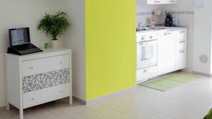 A modern living room with a flat-screen TV, a small white cabinet with a laptop, a green plant, and a kitchen area with white cabinets and appliances. - Zona Fiera Rho Però, IRCCS Ospedale Galeazzi - Sant'Ambrogio for families  (Rho)