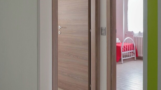 A hallway with a wooden door leading to a room with a white radiator and a red bedspread. - Zona Fiera Rho Però, IRCCS Ospedale Galeazzi - Sant'Ambrogio for families  (Rho)