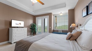 Un dormitorio con cama, ventilador de techo, televisor, cómoda, ventana con persianas y una planta.