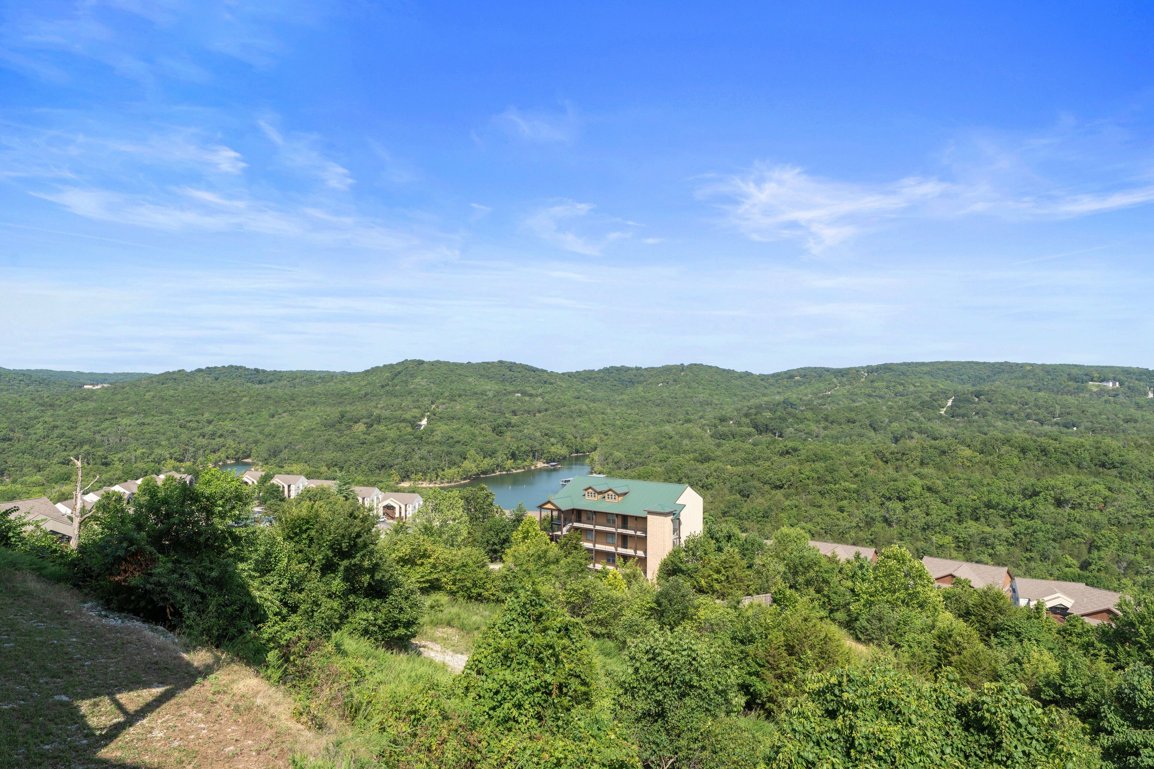 A multi-story building with a green roof, surrounded by dense greenery and hills in the background.