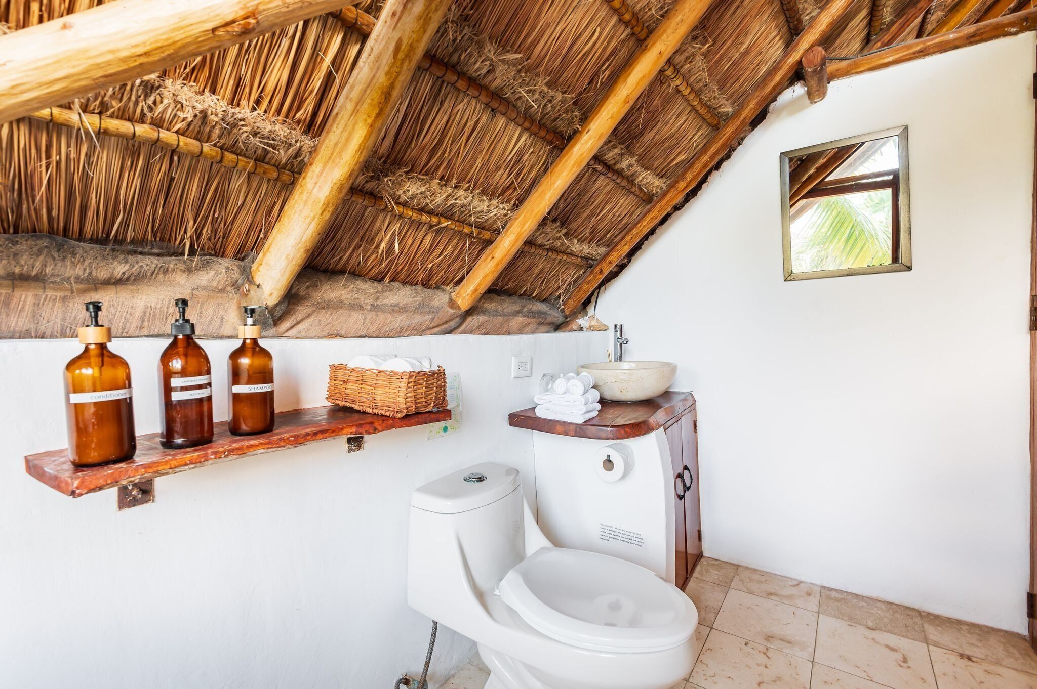Een badkamer met een rustieke inrichting, voorzien van een rieten dak, een wit toilet, een houten plankje met toiletartikelen en een klein raam.
