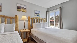 A hotel room with two beds, a wooden headboard, a nightstand with a lamp, and a window with a view of a mountainous landscape.