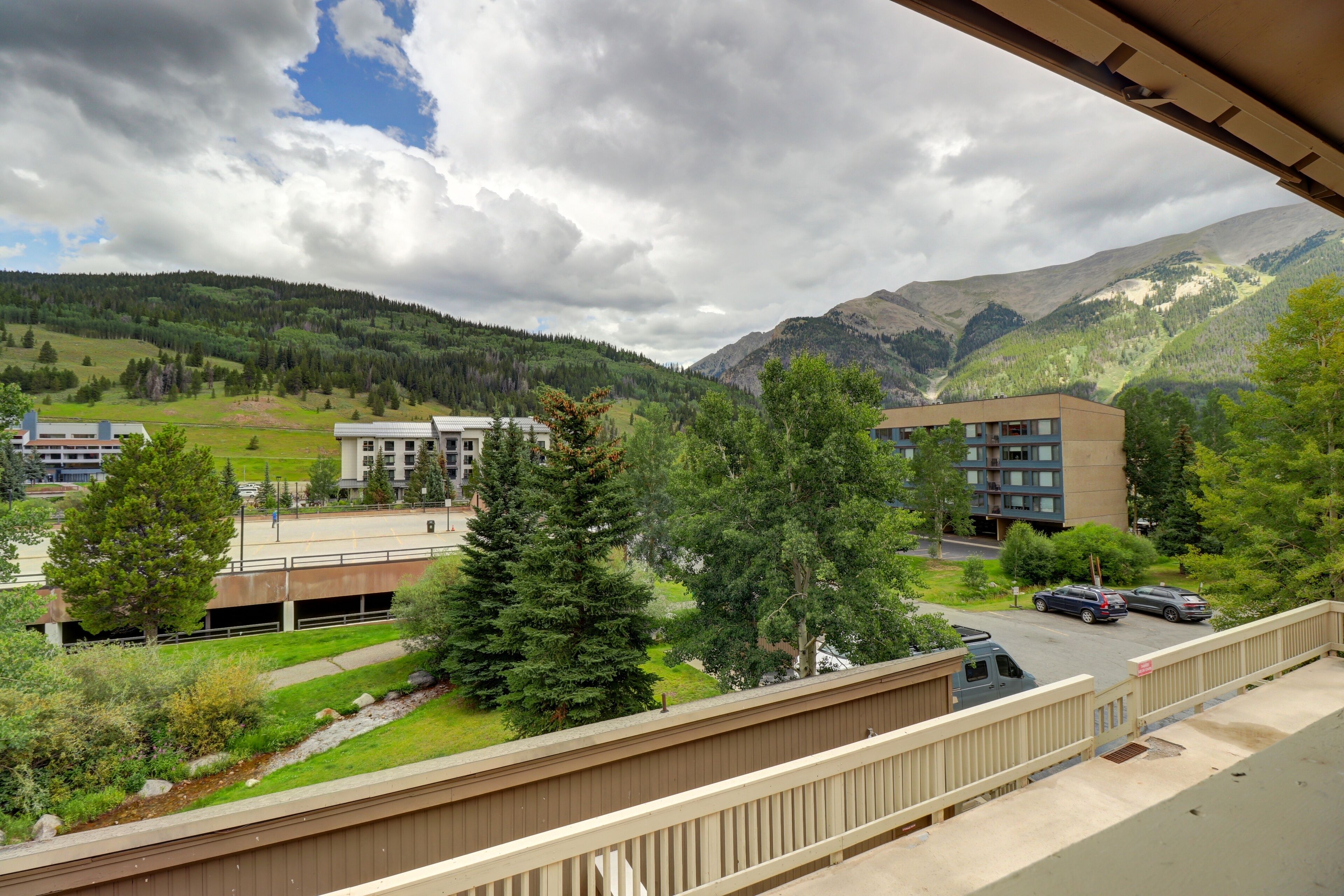 Une vue depuis un balcon donnant sur un parking où se trouvent des voitures, un immeuble et un paysage montagneux.