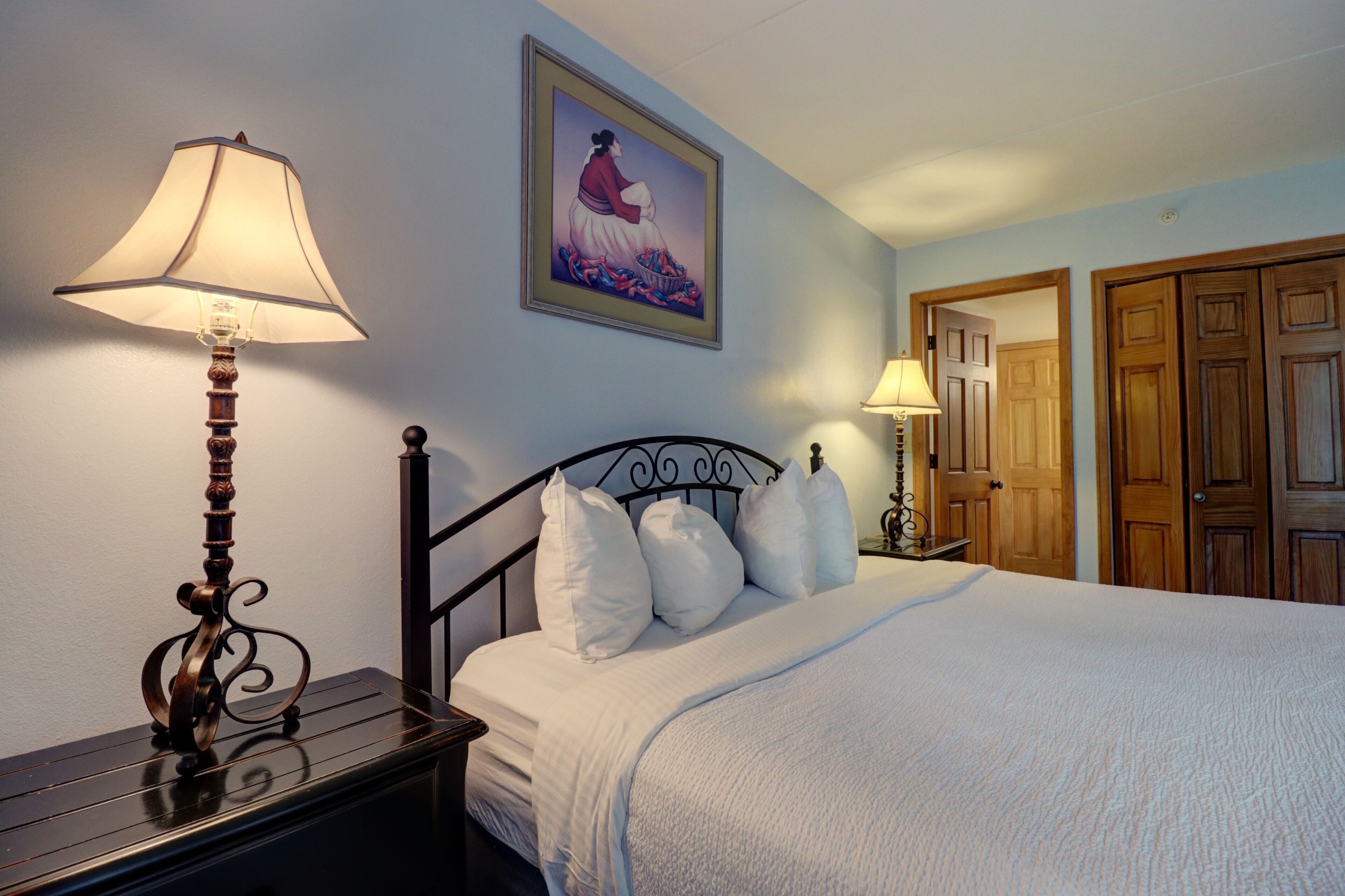 Una cama bien hecha con sábanas y almohadas blancas, una mesita de noche de madera con una lámpara y un cuadro en la pared.