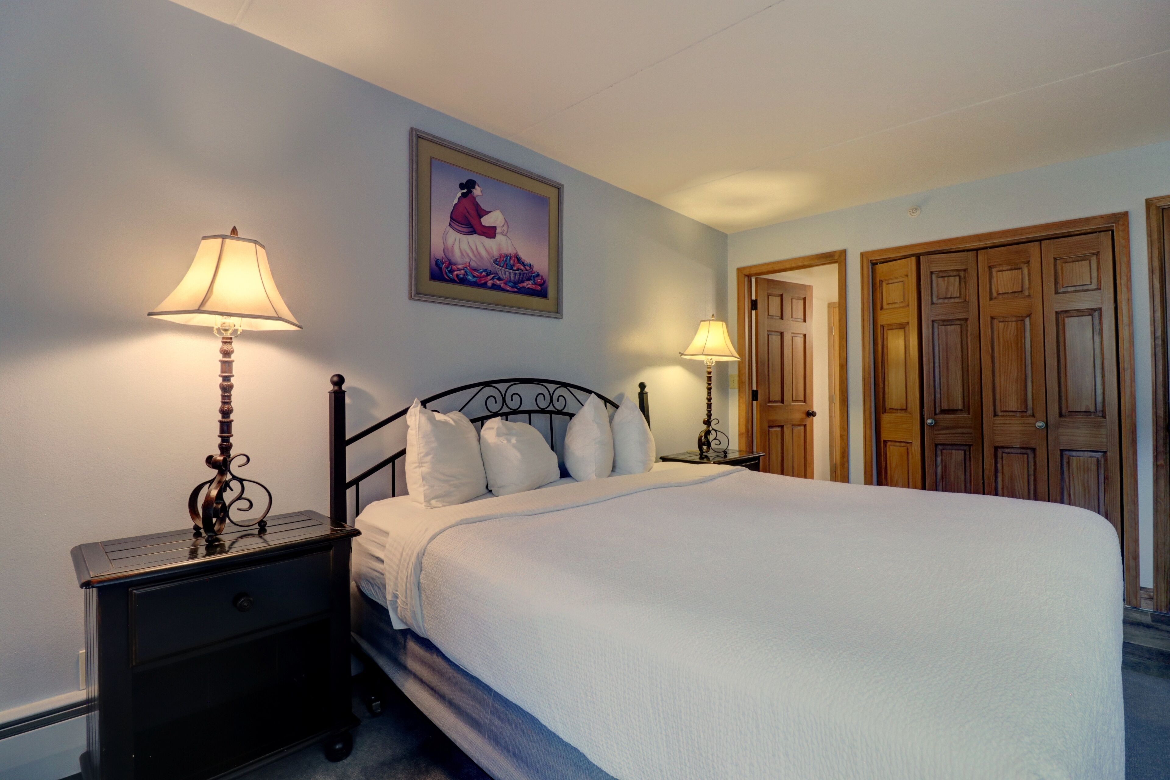 A neatly made bed with white linens, two bedside tables with lamps, a framed picture on the wall, and a set of wooden doors.