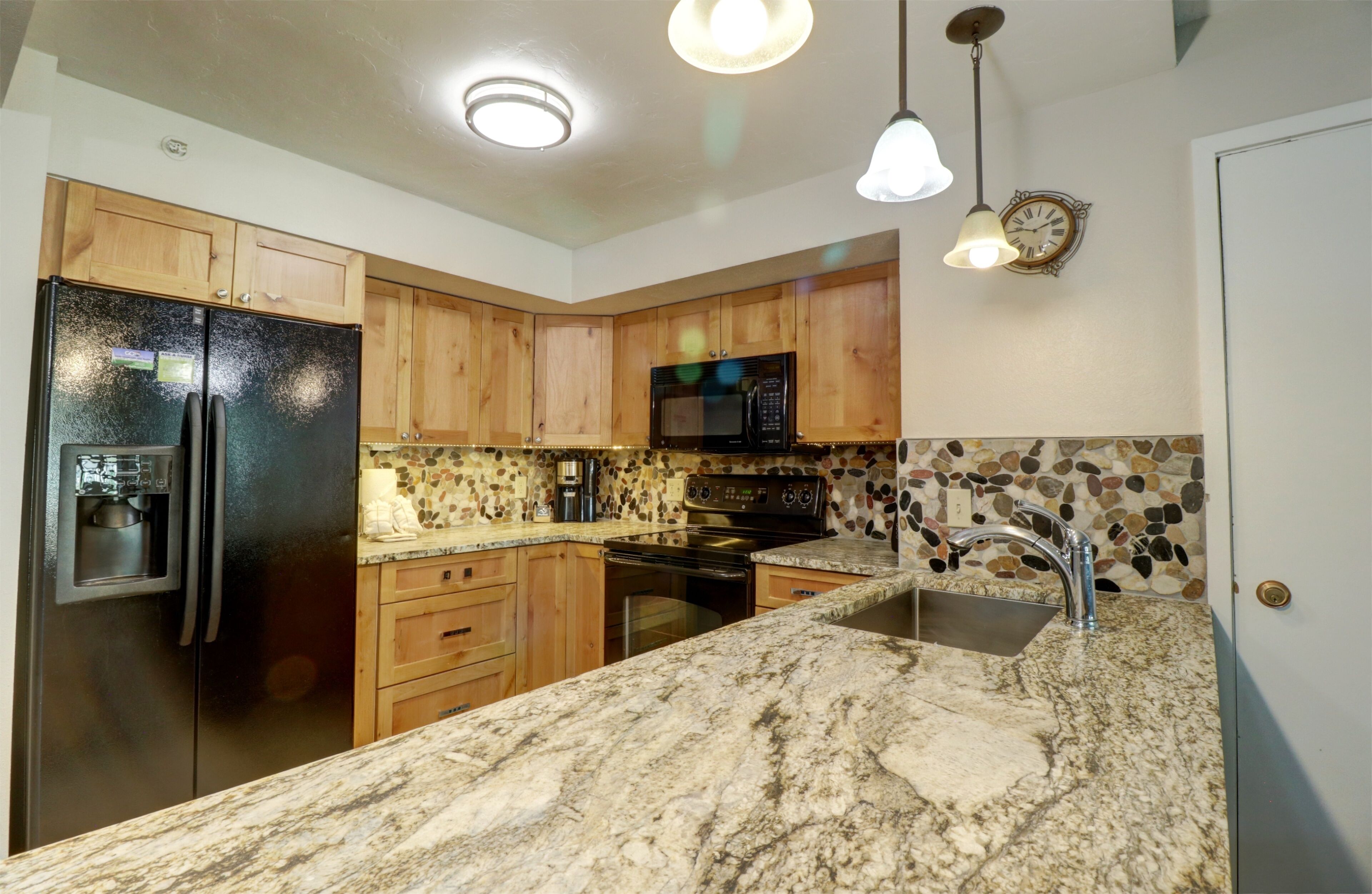 A kitchen with wooden cabinets, a black refrigerator, a microwave, an oven, granite countertops, and a stone backsplash.