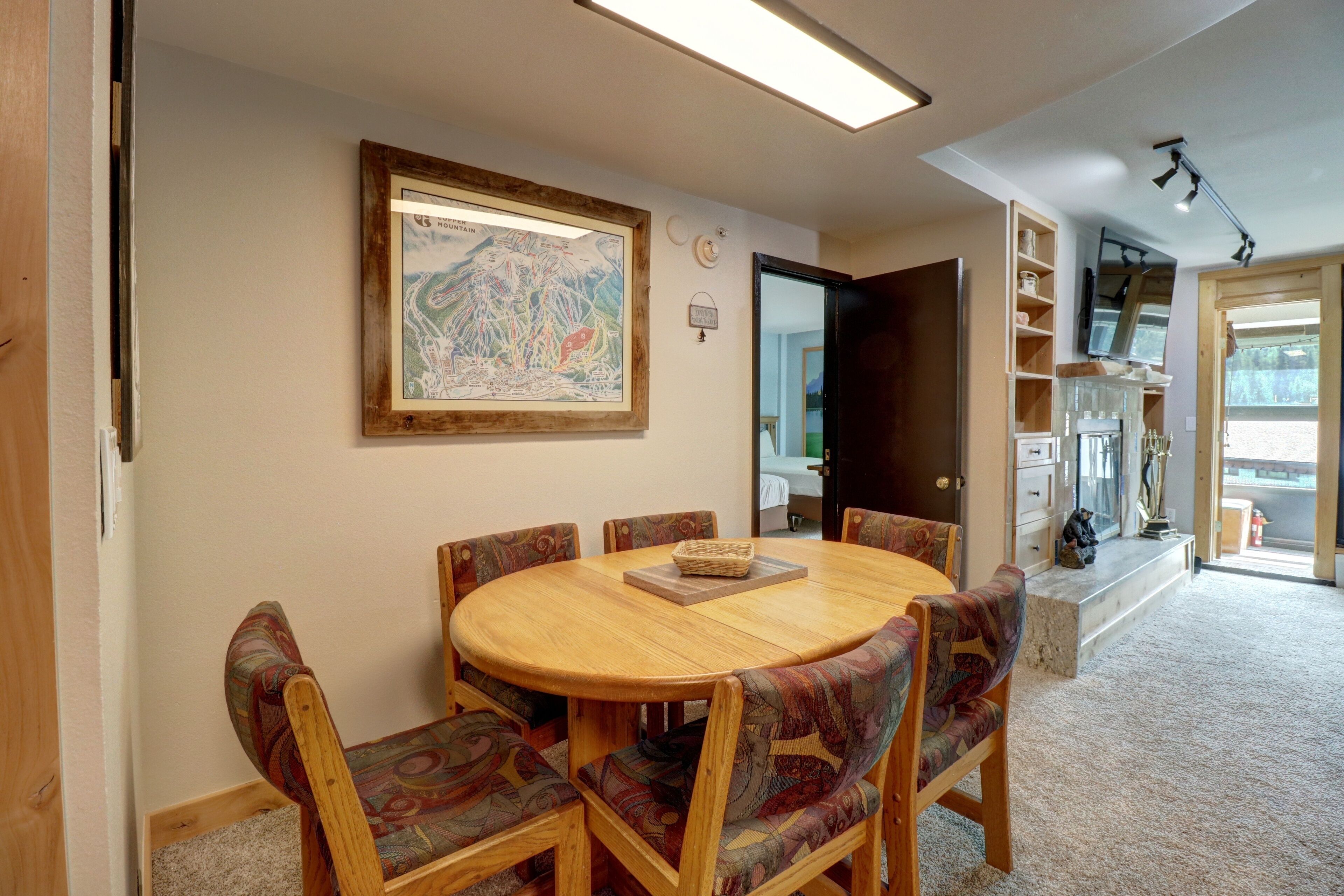 A dining area with a round wooden table and patterned chairs, a framed map on the wall, and a view of the kitchen and living area through an open door.