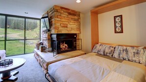 A bedroom with a stone fireplace, a large bed, a view of a forest through a window, and a framed picture on the wall.