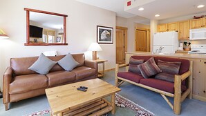 A living room with a brown leather sofa, a wooden coffee table, a red sofa, a wooden cabinet, and a kitchen area with wooden cabinets and a white refrigerator. - Tx314 Taylors Crossing 2 Bedroom Condo (Copper Mountain)