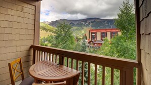A balcony with a wooden table and chair, overlooking a mountainous landscape with a lake and buildings. - Tx314 Taylors Crossing 2 Bedroom Condo (Copper Mountain)