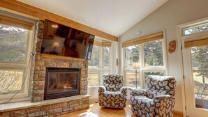 A cozy living room with a stone fireplace, large windows, and two patterned armchairs.