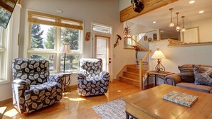 A spacious living room with two patterned armchairs, a wooden coffee table, and a sofa. A staircase with wooden railings leads to an upper level.