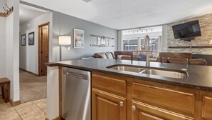 A modern kitchen with a central island, stainless steel sink, wooden cabinets, and a built-in TV. - Be306b Bridge End 1br 1ba 2 Bedroom Condo (Copper Mountain)