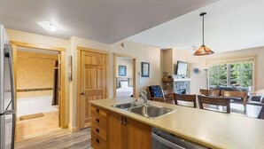 A kitchen with wooden cabinets, a stainless steel sink, and a dishwasher. There is a dining area with a table and chairs. - Pp219 Passage Point 2br 2ba 2 Bedroom Condo (Copper Mountain)