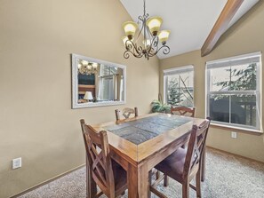 A dining room with a wooden table and chairs, a chandelier, a mirror, and windows with a view of trees.