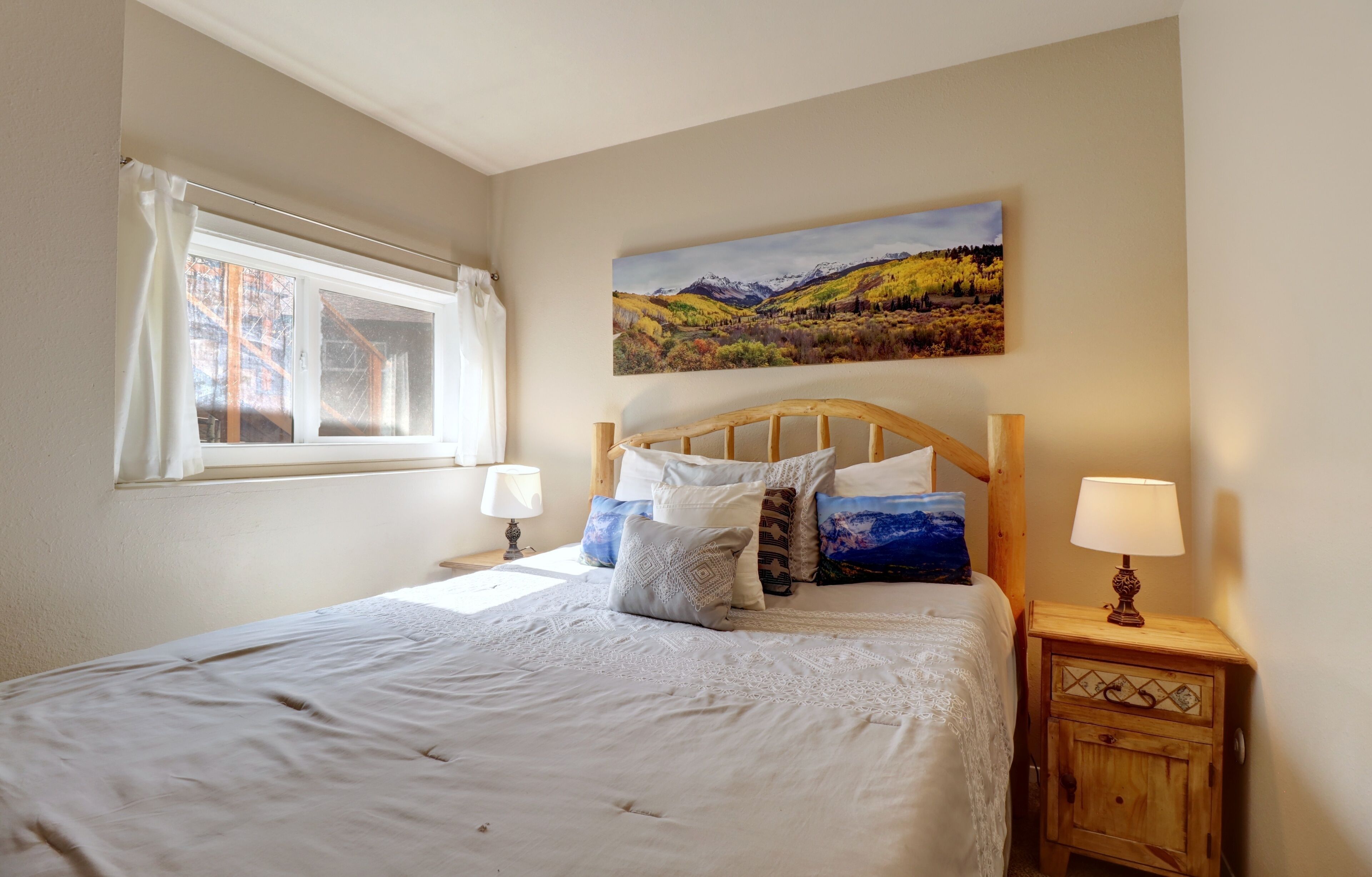 Een slaapkamer met een houten hoofdbord, een groot landschap schilderij, twee bedlampen en een raam met gordijnen.
