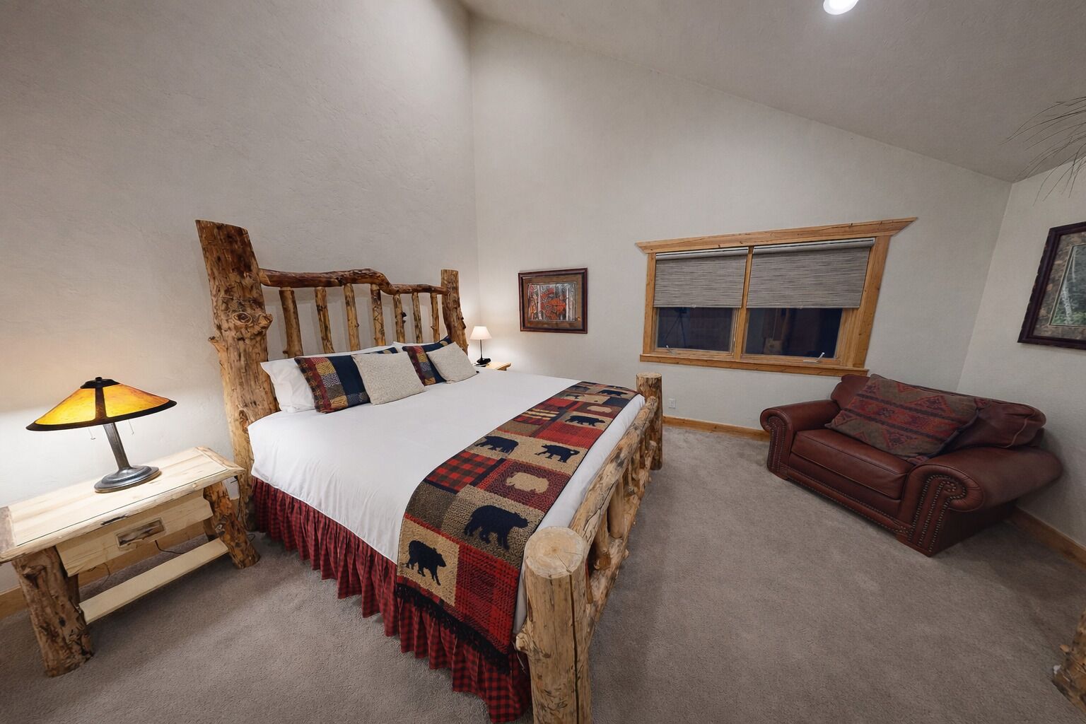 Un dormitorio con estructura de cama de madera, una mesita de noche con lámpara, una ventana con persianas y un sofá de cuero marrón.
