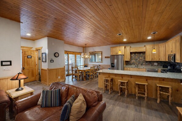 Un amplio salón con un sofá de cuero marrón, una mesa de comedor de madera y una cocina con armarios de madera y encimera de granito.