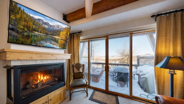 A cozy living room with a fireplace, a large window with a view of a snowy deck, and a mounted painting of a forest scene.