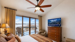 A hotel room with a large bed, a ceiling fan, a television displaying a news channel, and a view of a snowy landscape through the windows.