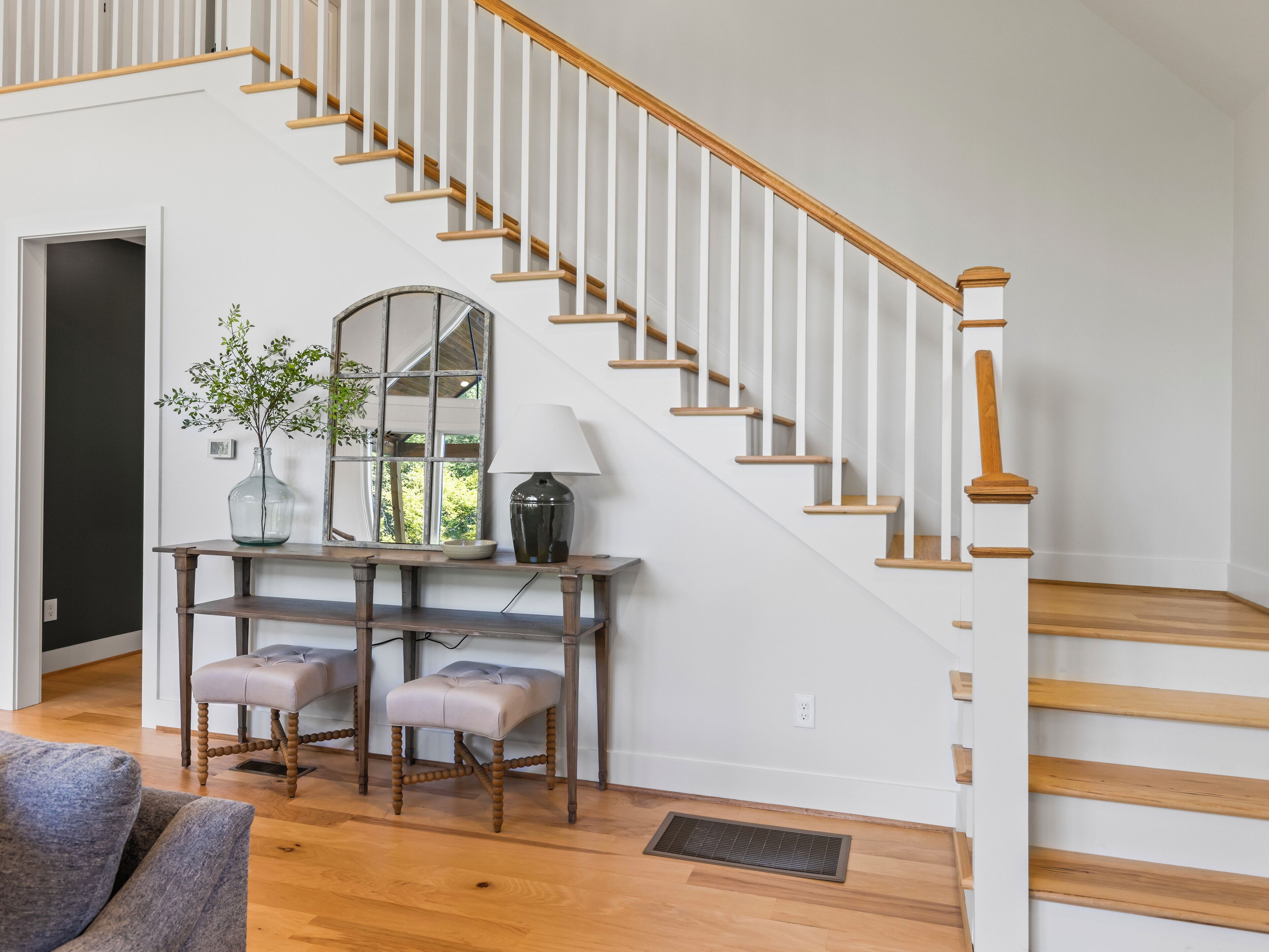 A staircase with white railings and wooden steps, leading to a landing with a mirror, a vase with green plants, and a table with two stools.