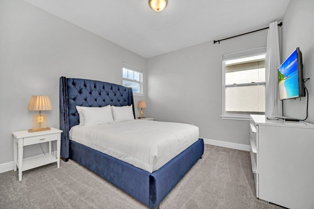 A bedroom with a tufted headboard, a white bedspread, a bedside table with a lamp, a dresser, and a TV mounted on the wall.