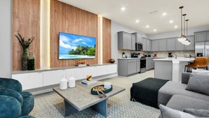 A modern living room with a flat-screen TV, a wooden accent wall, a kitchen with grey cabinets, and a dining area with a bar.