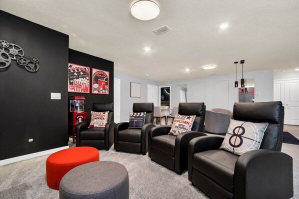 Une salle de cinéma privée avec des fauteuils inclinables en cuir noir, un pouf rouge et des affiches de films accrochées au mur.