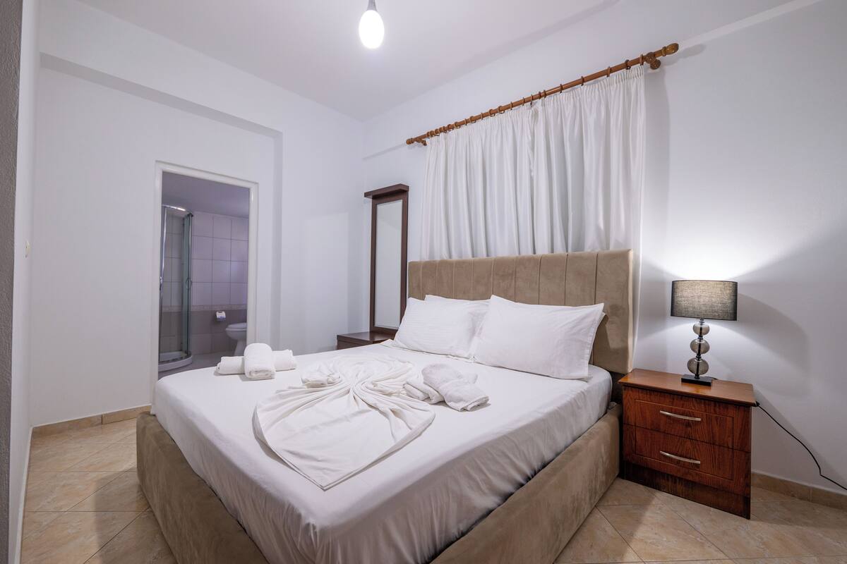 A neatly made bed with white linens and towels, a wooden nightstand with a lamp, and a bathroom with a shower and toilet visible.