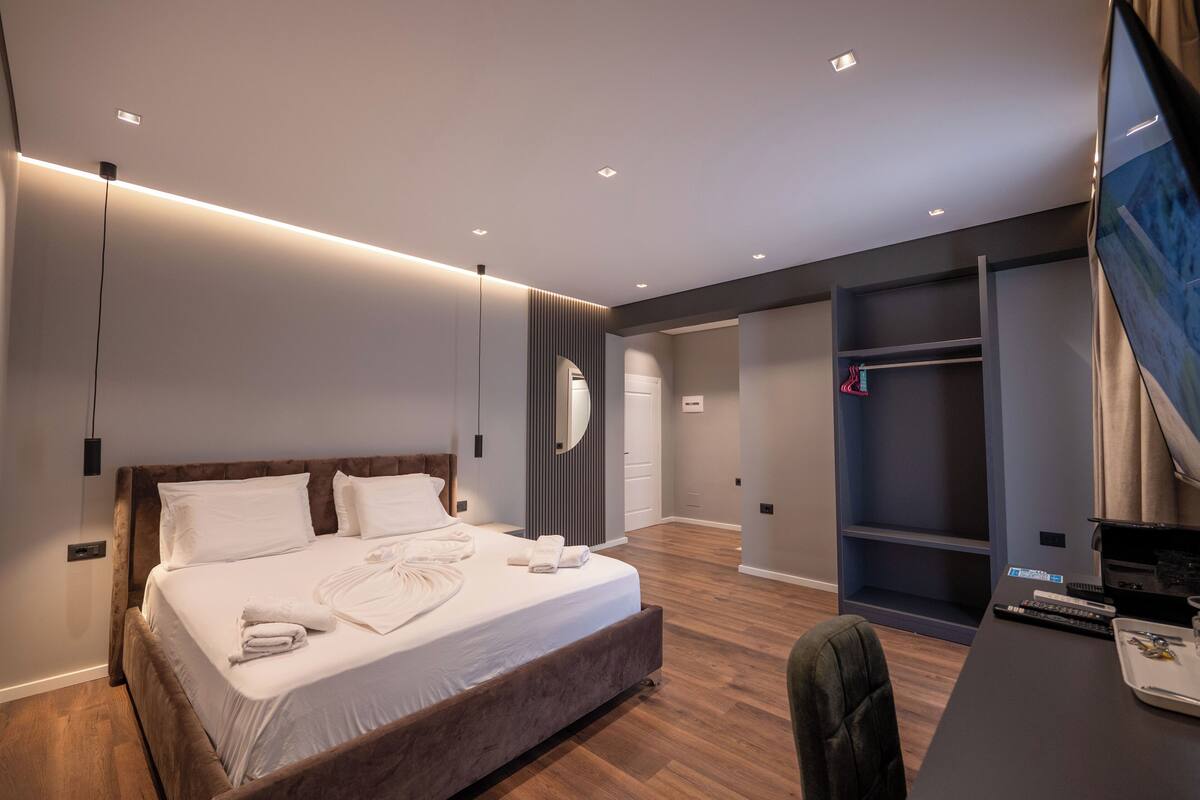 A modern hotel room with a large bed, a desk with a laptop, and a wardrobe.