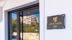 Una entrada de hotel con puerta de cristal, un letrero que indica "HOTEL ERIONI 49", y vista de la calle y edificios al fondo.