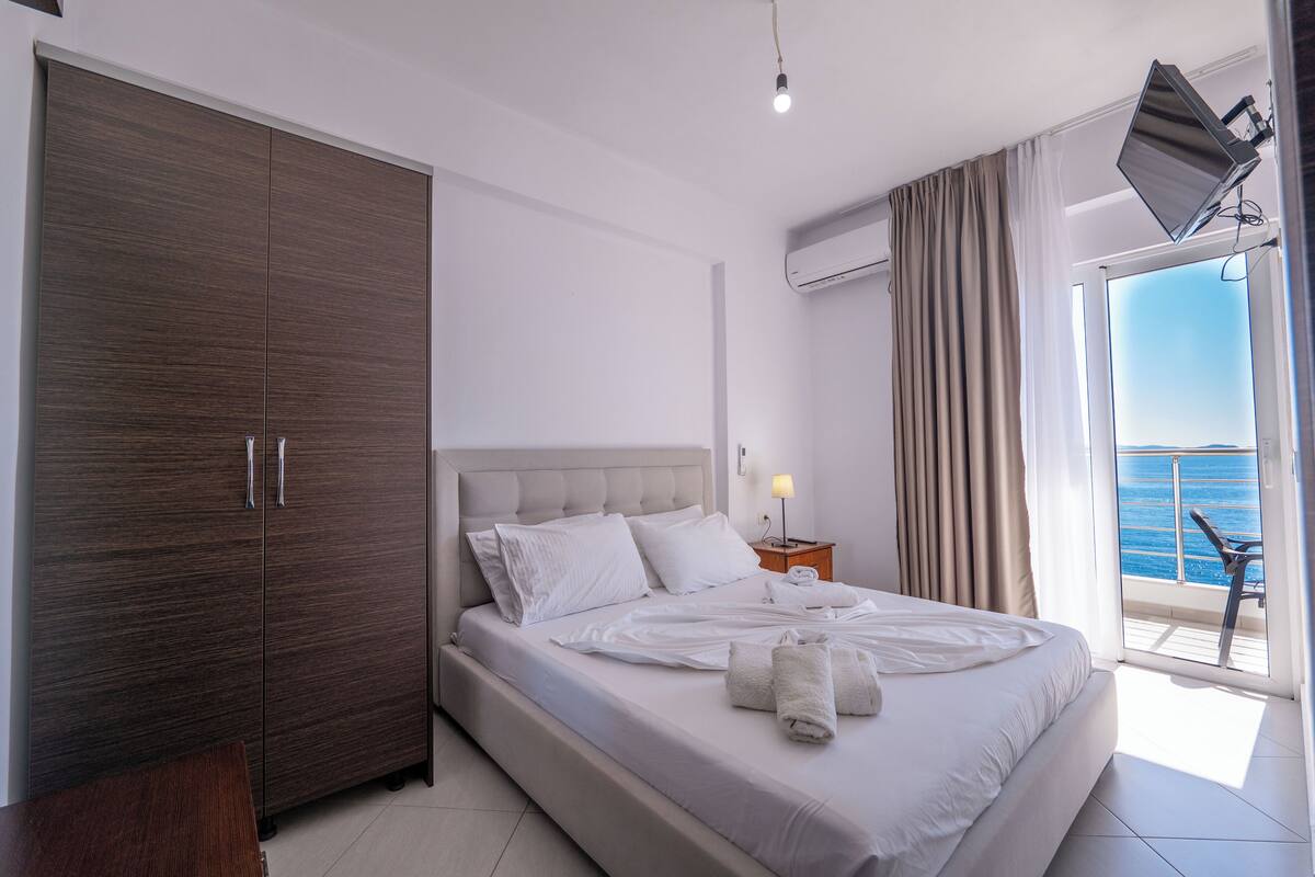 A modern hotel room with a large bed, a wardrobe, a nightstand with a lamp, and a balcony with a view of the sea.