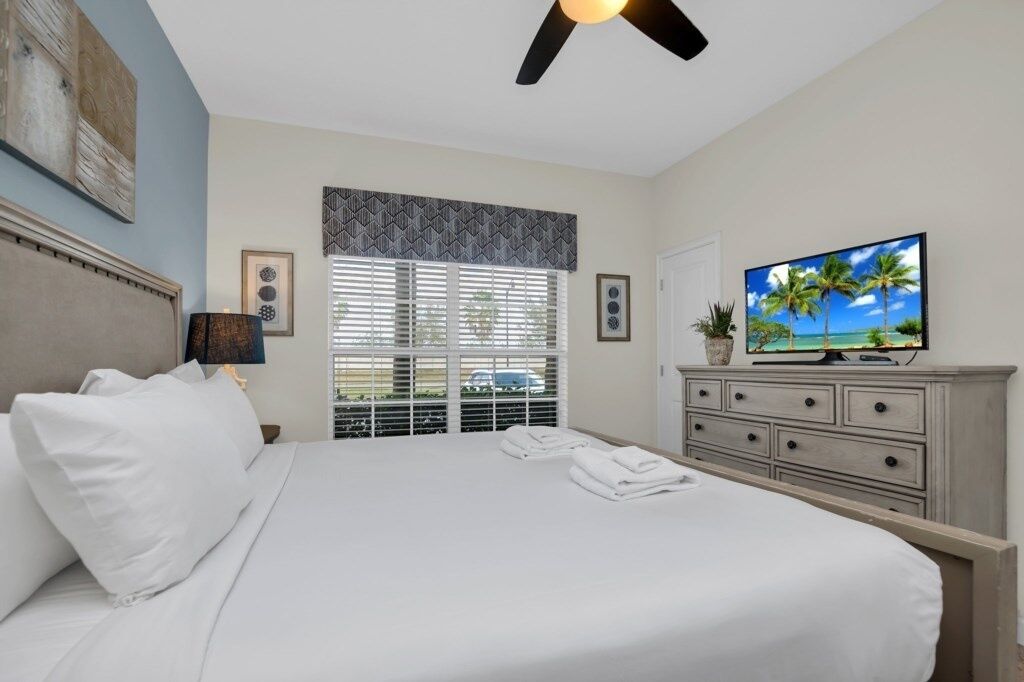Een slaapkamer met een bed, een plafondventilator, een televisie en een raam met jaloezieën.
