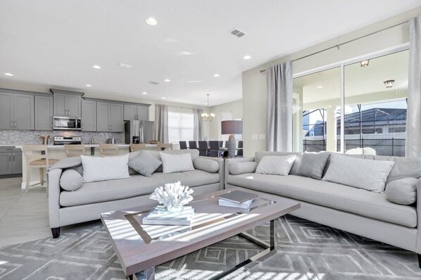 A modern living room with a sectional sofa, a coffee table, and a kitchen in the background. - Florida Garden Luxury Home With Private Pool & Spa 6 Bedroom Villa (Kissimmee)