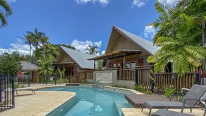 A swimming pool with lounge chairs, surrounded by wooden buildings with thatched roofs, palm trees, and a clear blue sky. - Pure Magnetic Villa 10 (Nelly Bay)