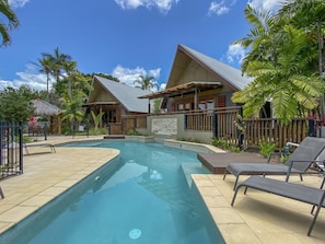 A swimming pool with lounge chairs, surrounded by wooden buildings with thatched roofs, palm trees, and a clear blue sky.