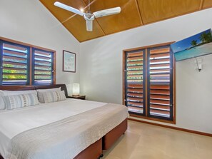 A bedroom with a bed, a ceiling fan, and two windows with shutters.