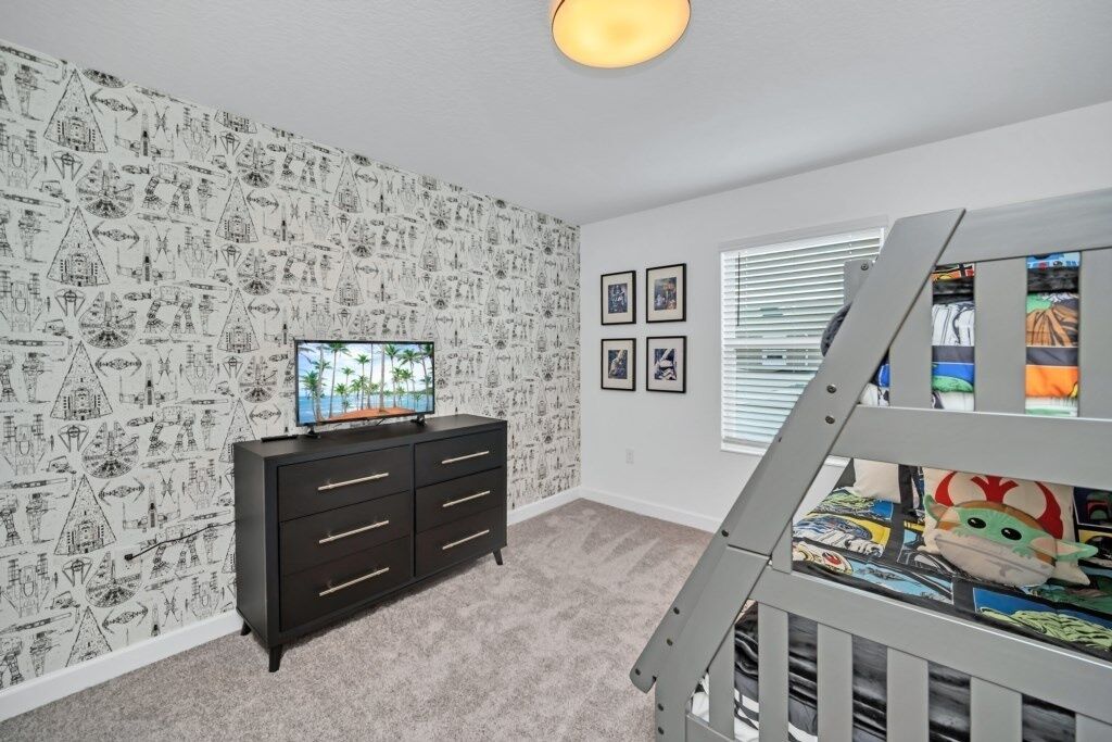 A room with a patterned wallpaper, a TV on a black dresser, a window with blinds, and a crib with a colorful blanket.