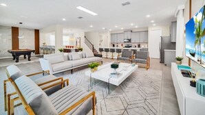 Ein modernes Wohnzimmer mit Sofa, Couchtisch und Sesseln, ein Küchenbereich mit Schränken und Geräten sowie ein Billardtisch.